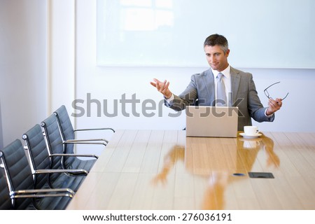Business manager sitting at the end of a meeting table, with cup of cafe and laptop, moving hands with lecturer attitudes toward empty chairs.