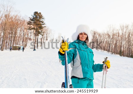Attractive young female on a ski resort. Friendly smiling young woman wears fur hat and ski clothes. Hands poles and skis. Standing back to slope and forest. Half-length portrait