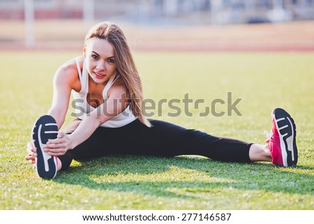 Sport and stretching on a field. Young attractive long haired woman in sportswear holds her foot to stretch well. Girl sitting on a grass while stretching. Bright and warm scene
