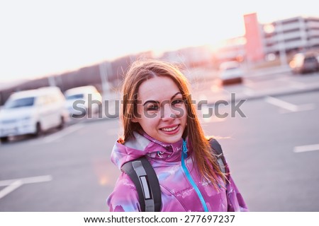 Attractive smiling young female with backpack straps on her shoulders. Stands on a parking lot in urban scenery at sunset. Looking right to camera