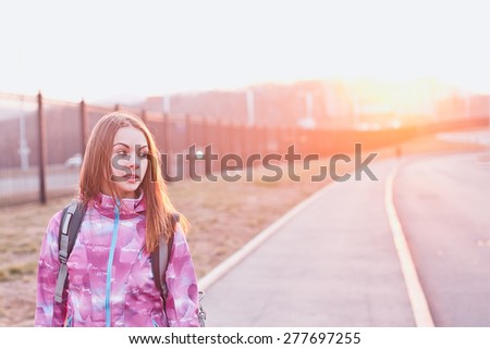Attractive girl in windbreaker with backpack looking aside on a long valley at a sunset. Urban scenery. Traveling, life and freedom concept. Copy space