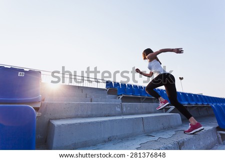 Sporty girl in white shirt running fast to a sunset on a stadium. Side view. Dynamic photo. Young woman in sportswear training on a stadium. Copy space. Sport lifestyle concept