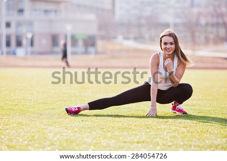 Young woman in sportswear with long hair stretching on a field in a campus. Sit with straight leg and hand raised to a chin. Sunny and bright photo. Concept of healthy lifestyle