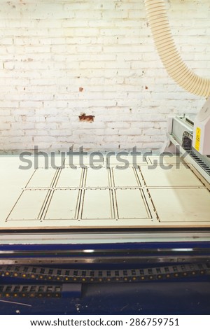 Working table of CNC wood router machine, white painted brick wall with a hole and flexible ventilation duct pipe from a machine. Router table with etching on product deck and belt drive above it