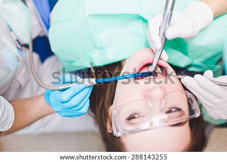 Young female patient with dental cases in dentist chair looking at a dentist assistant during treatment. Hands with dental instruments dental mirror and dental drill in patients mouth. Plan view