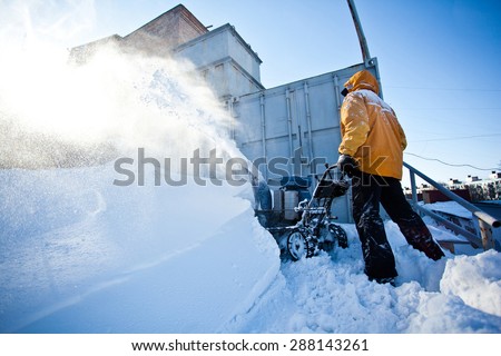 Worker in yellow winter jacket removes snow near his garage with a snow throwing machine at a sunny winter day. Clouds of snow dust around. Seasonal photo