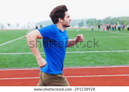 Attractive man runner in blue shirt running fast in front of football field with activity on background