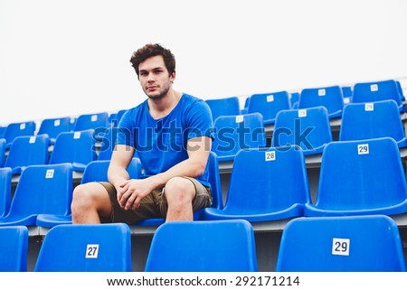 Attractive sporty young man model in blue shirt relaxing on blue stadium seats after training staring into camera. Copy space