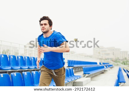 Attractive male model in blue shirt running on a stadium between blue seats. Sport and healthy lifestyle in a city concept. Runner in blue shirt training on stadium in urban scenery
