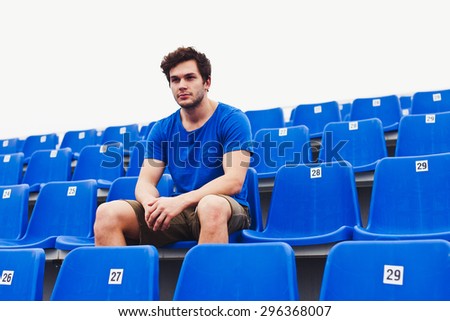 Attractive sporty young man model in blue shirt relaxing on blue stadium seats after training staring at field. Copy space