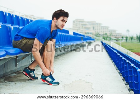 Young attractive male model in blue shirt tying laces on his sports shoes and looking at field before run on a stadium stands after workout. Copy space