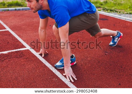 Man runner in blue shirt and shorts and sport shoes in steady position before run at start of race track preparing for run on a stadium