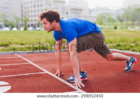 Man runner in blue shirt and shorts and sport shoes in steady position before run at start of race track preparing for run on a stadium perspective view with university campus on background