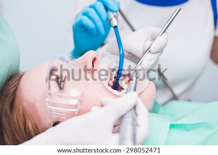 Girl patient with protection glasses with dental braces and mouth retractor during treatment at dentist office with dentist and assistant with saliva ejector and dental mirror dentist tools side view