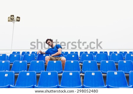 Attractive sporty young man model in blue shirt sitting on blue stadium seats relaxing after training staring at field. Straight view on stadium chairs. Toned whites. Copy space