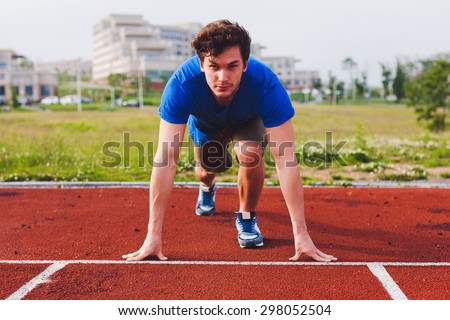 Man runner in blue shirt and shorts and sport shoes in steady position before run at start of race track preparing for run on a stadium with university campus on background