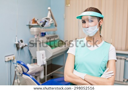 Young beautiful woman dentist in protective mask and full face shield standing in her office in front of dentist chair with hands crossed