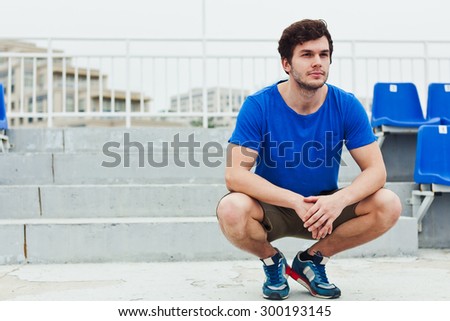 Young attractive man model sitting in squats at stadium stairs after training looking aside relax in casual pose copy space