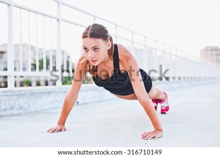Young attractive girl athlete in sportswear performing push-ups on stadium on concrete floor. Wide hands position on photo with stressed look. Torso, chest, shoulders and hands affecting exercise