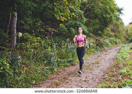 Young beautiful sporty girl training on country road near green forest during summer autumn season with lots of leaves fallen