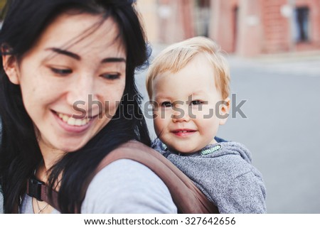 Beautiful young asian woman with freckles and her son relax outdoors. Mother brunette with dark hair and her son is blond. Unusual appearance and heredity concept. Baby sits in sling on mothers back