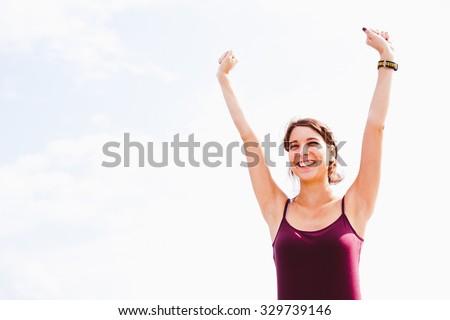 Beautiful sporty young woman rising hands up after training with wearable smart watch on her wrist. Copy space with bright sky on background
