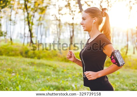 Young attractive female runner with armband running on sunset in park with earphones listening to music during training. Copy space and sun on background