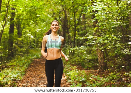 Young beautiful sporty girl in short green sports top training in green forest during summer autumn season with lots of leaves fallen. Front view with copy space