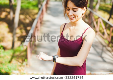Young athletic girl runner in sportswear checking out and adjusting her stopwatch cardio monitor wearable smart watch on hand after training in a city park. Concept of wearable devices. Copy space