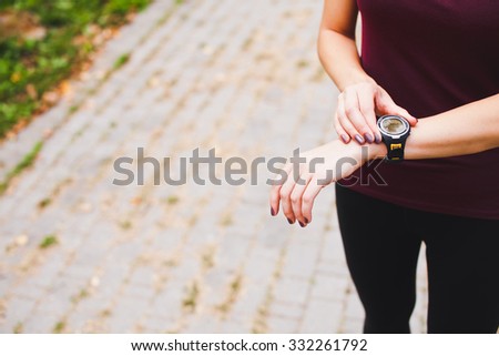 Young athletic girl runner in sportswear checking out and adjusting her stopwatch cardio monitor wearable smart watch on hand after training in a city park. Concept of wearable devices. Copy space