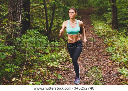 Young beautiful sporty girl in short green sports top training in green forest during summer autumn season with lots of fallen leaves on path. Front view with copy space full length portrait of runner