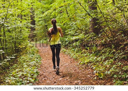 Young athletic sporty girl with long hair in green sleeveless shirt training in green forest during summer autumn season with lots of leaves fallen on forest path. Back view with copy space