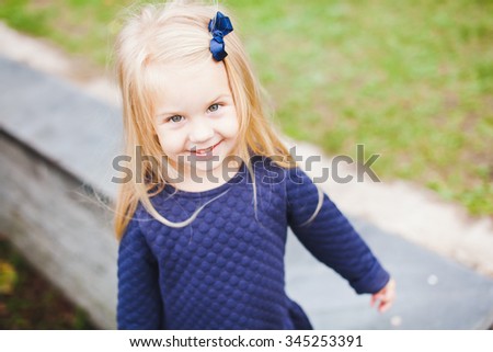 Little beautiful blonde girl with small blue bow in her hair wearing blue dress in park during walk on sunny summer autumn day. Young beautiful kid smiling and posing. Copy space
