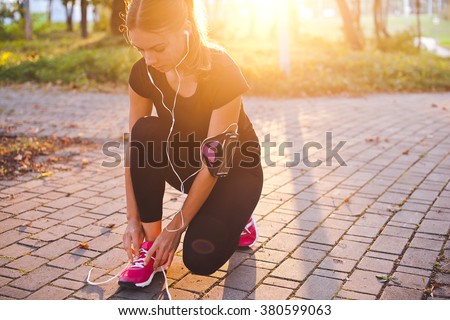 Young beautiful girl athlete with armband and earphones listening to music and tying laces during training in park with sunset and sunbeam on background. Copy space. Bright and warm photo