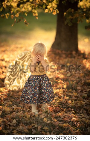 Adorable toddler girl staying under the tree in the park. Hiding your face. Arms on her face. Autumn. Colorful leaves. Magic back light. Sunset.