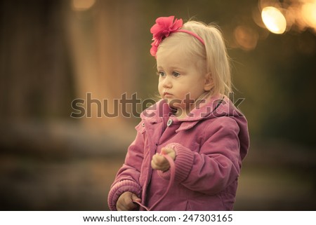 Little sad girl in the park. Pink flower in her hair. Pink coat. Art blurry background. Natural light. Sunset.