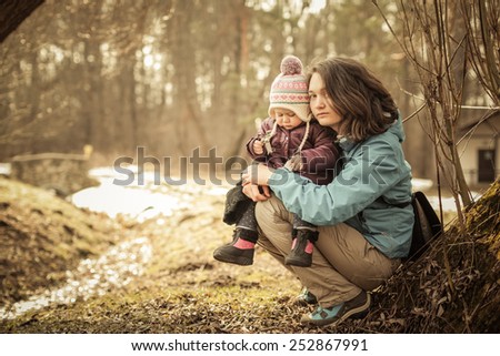 Adorable toddler girl with Mom sitting on the ground in the park. Cold spring. Shiny stream background. Forest without leaves.