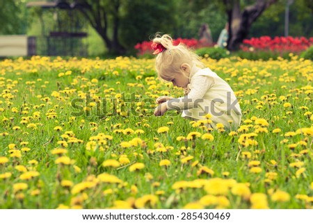 Little girl playing in the dandelion field. White dress with light brown bow. Red ribbon on her head. Red flowers background.