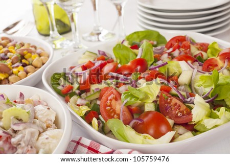 An assortment of salads on a buffet table. Potato salad, bean salad and fresh mixed salad arranged on a white table with glasses, cutlery and plates. Focus on fresh mixed salad.