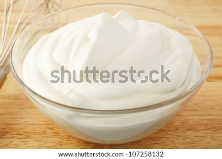 A bowl of softly whipped cream in a glass bowl, on a wooden board, whisk in background.