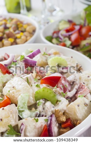 An assortment of salads on a buffet table. Potato salad, bean salad and fresh mixed salad arranged on a white table with glasses, cutlery and plates. Focus on potato salad.