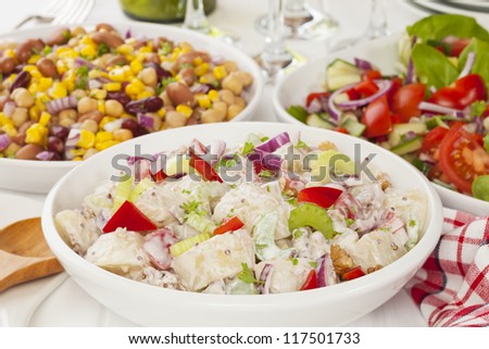 An assortment of salads on a buffet table. Potato salad, bean salad and fresh mixed salad arranged on a white table with glasses, cutlery and plates.