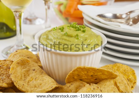 Guacamole topped with fresh green chilli, with corn chips for dipping, set out on a buffet table.