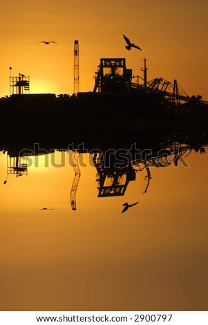 Port at sunset, ship and shiploader are silhouetted against orange sky with a couple of seagulls flying through.