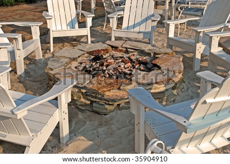 white wooden chairs form a circle around a stone firepit on sand