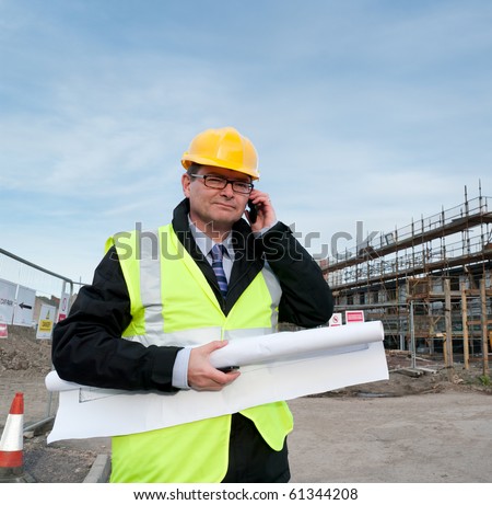 Architect or engineer at work on a building site. Holding plans for construction work. Confident gaze and smile at camera. Using telephone.
