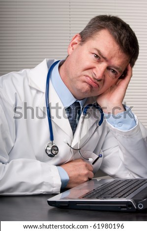 Physician at desk in surgery leans on hand and looks unhappy and bored. Looking  into distance. Humor.