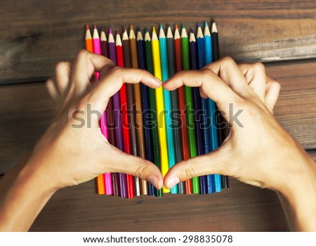 Female hands in the shape of a heart on colored pencils on a background of dark wood tables