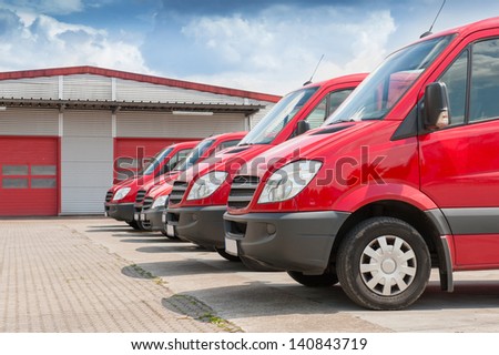 Row Of Red Delivery And Service Van, Trucks And Cars In Front Of A Factory And Warehouse Distribution Plant