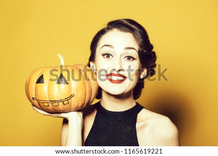 Smiling woman with beautiful face and retro hairdo and red lips holding pumpkin in studio on yellow background. Halloween concept